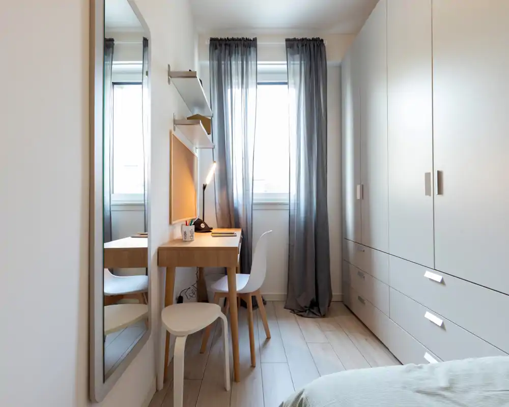 Bright, minimalist bedroom corner with a compact wooden desk, chair and wardrobe; soft natural light filters through sheer curtains and a bedside/desk lamp adds warmth — ideal for a small, well-organized sleeping space.