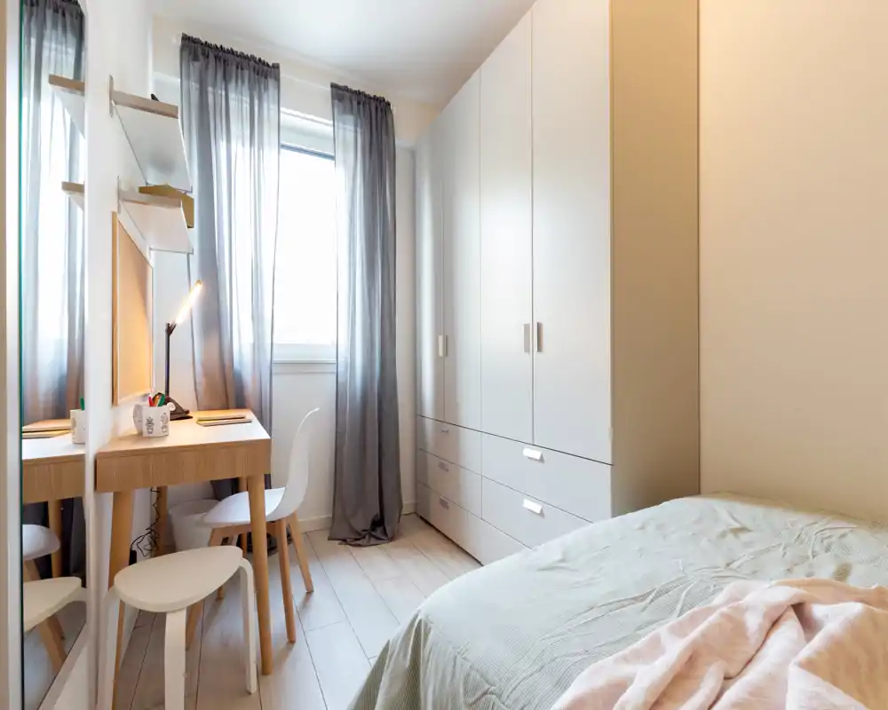 Bright, compact bedroom with a visible double bed, large wardrobe and a small desk by the window — modern, minimalist styling with natural light.