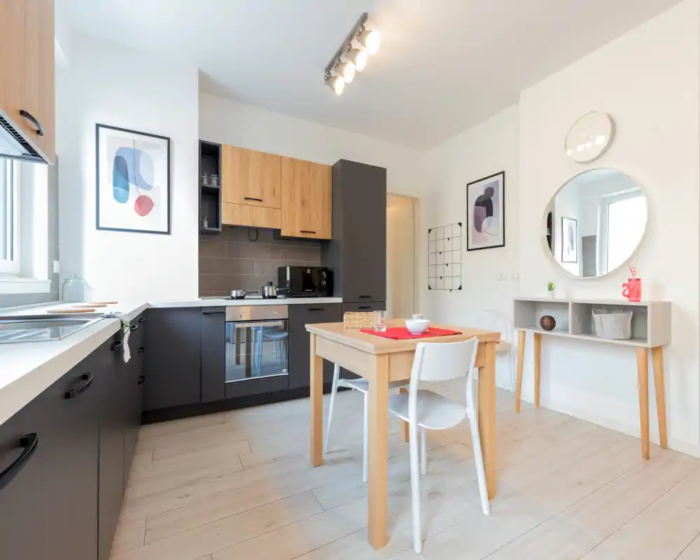 Bright modern kitchen with matte gray lower cabinets, wood upper cabinets, integrated oven and a small wooden dining table set for two. Clean, minimal décor with wall art and a round mirror brighten the space.