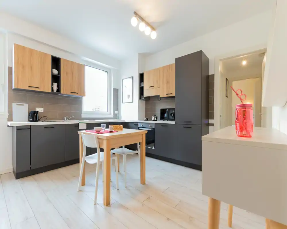 Bright, modern kitchen with matte dark lower cabinets and light wood upper cabinets, a small dining table in the center and natural light from a large window.