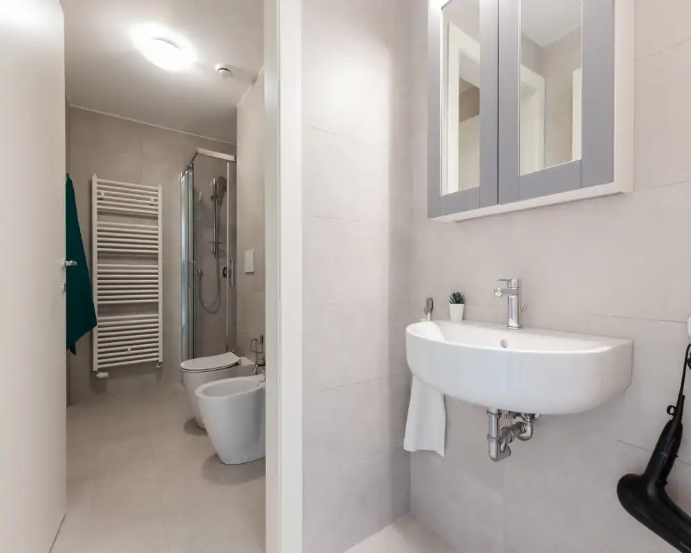 Modern, compact bathroom with wall-mounted sink, mirrored cabinet, toilet, bidet and a glass shower enclosure; light neutral tiles and good lighting create a clean, contemporary feel.