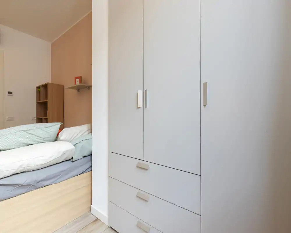 Coin chambre moderne et confortable avec un lit simple visible et une grande armoire blanche. Literie douce et tons neutres pour une atmosphère calme, idéale pour une annonce locative.