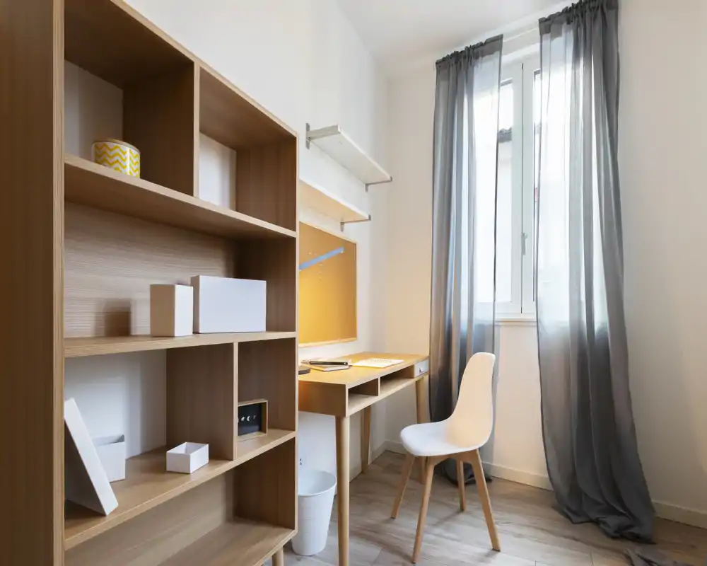 Bright, compact home office with a wooden desk, shelving unit and a comfortable chair by the window providing natural light — ideal for focused work or study.