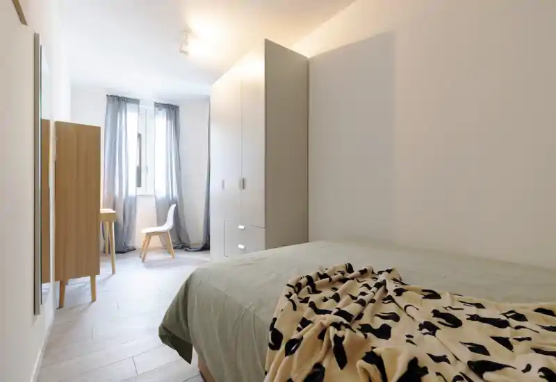 Bright, minimal bedroom with a neatly made bed, a wardrobe and a small desk by the window. Soft natural light and neutral tones create a calm, welcoming atmosphere ideal for listings.