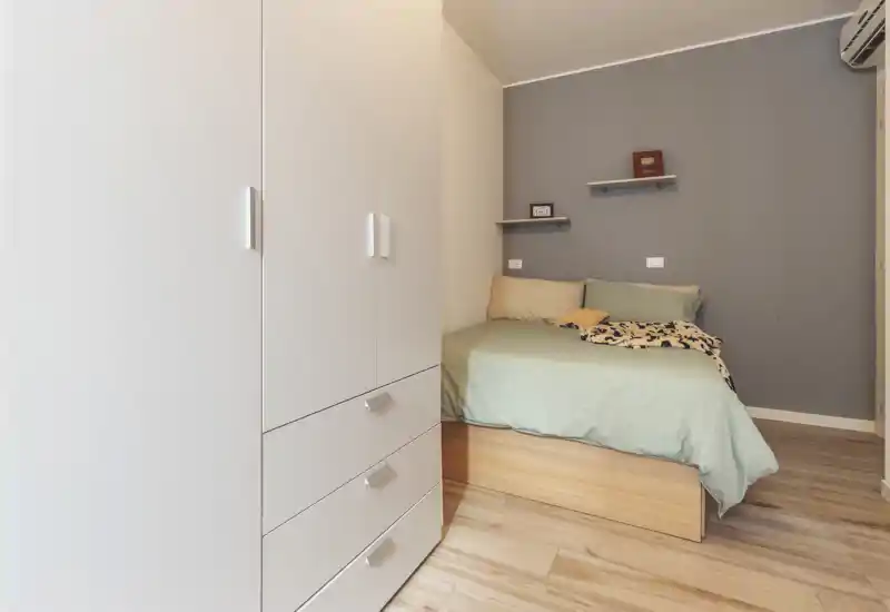 Compact, well-lit bedroom with a double bed dressed in muted green linens, soft pillows and a small wooden base. A white wardrobe in the foreground and minimalist shelves on a grey accent wall create a clean, modern look ideal for short-term rentals.