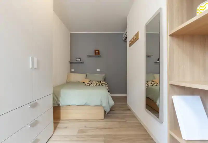 Cozy, well-lit single bedroom with a neatly made bed, soft green bedding, built-in storage and a full-length mirror — ideal for a compact rental or guest room.