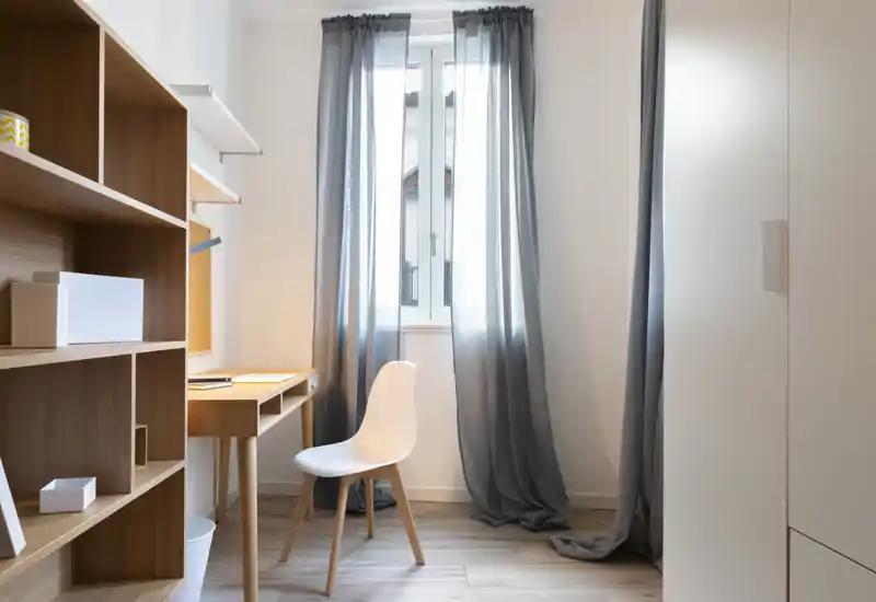 Bright compact bedroom with a small wooden desk and chair by the window, soft grey curtains and built-in storage creating a calm, functional space.