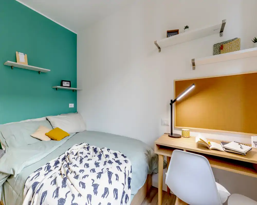 Camera da letto luminosa e accogliente con letto matrimoniale, biancheria a strati e una piccola scrivania con bacheca e lampada — perfetta per soggiorni confortevoli e smart working.
