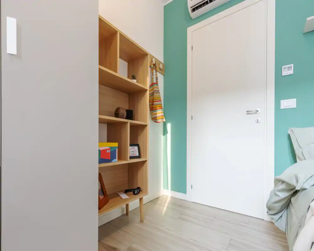 Angolo di camera da letto con letto parzialmente visibile a destra, una libreria in legno, parete verde pastello e porta bianca — ordinato, moderno e ben illuminato.