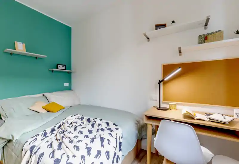 Camera da letto luminosa e accogliente con letto matrimoniale, biancheria a strati e una piccola scrivania con bacheca e lampada — perfetta per soggiorni confortevoli e smart working.