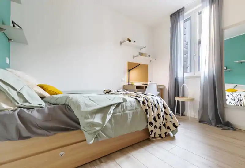 Camera da letto moderna e luminosa con letto ben rifatto, biancheria verde chiaro, plaid a fantasia e una scrivania vicino alla finestra — perfetta per annunci in affitto che valorizzano comfort e luce naturale.