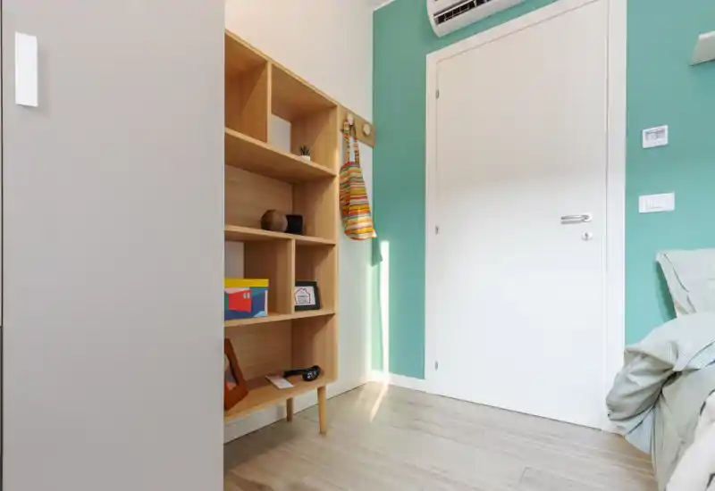 Angolo di camera da letto con letto parzialmente visibile a destra, una libreria in legno, parete verde pastello e porta bianca — ordinato, moderno e ben illuminato.