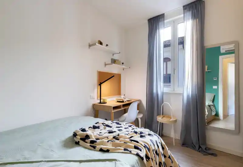 Camera da letto luminosa e ben arredata con letto ordinato, scrivania compatta in legno vicino alla finestra e luce naturale soffusa. Ideale per un soggiorno accogliente con area lavoro.