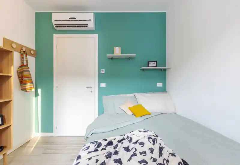 Camera da letto moderna e luminosa con parete accent color turchese, letto ben rifatto con cuscini decorativi, mensole minimal e climatizzatore.