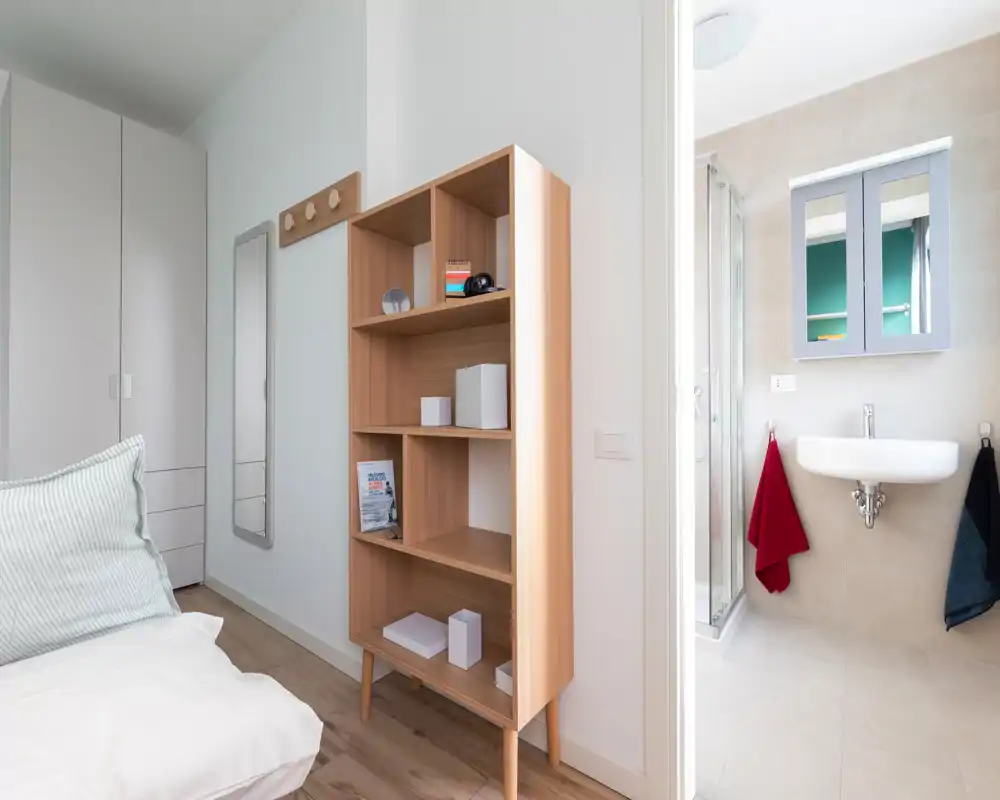 Bright, compact bedroom corner with a partially visible bed, wooden shelving unit and wardrobe; an en-suite bathroom is visible through an open door.