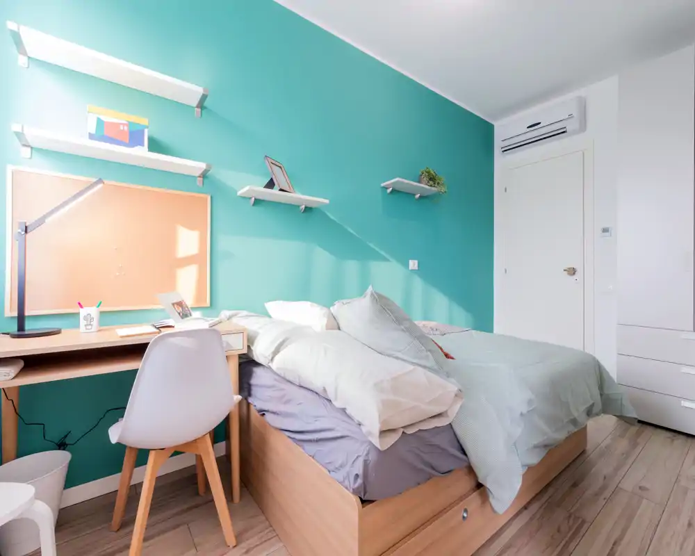 Bright, modern bedroom with a wooden bed, layered bedding and a small workspace. Teal accent wall and floating shelves give a fresh, cozy look ideal for rental photos.