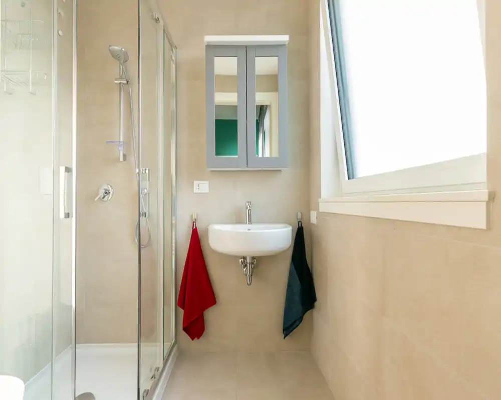 Bright, modern bathroom with a glass shower, wall-mounted sink and mirrored cabinet. Neutral tiles and a large window provide ample daylight.