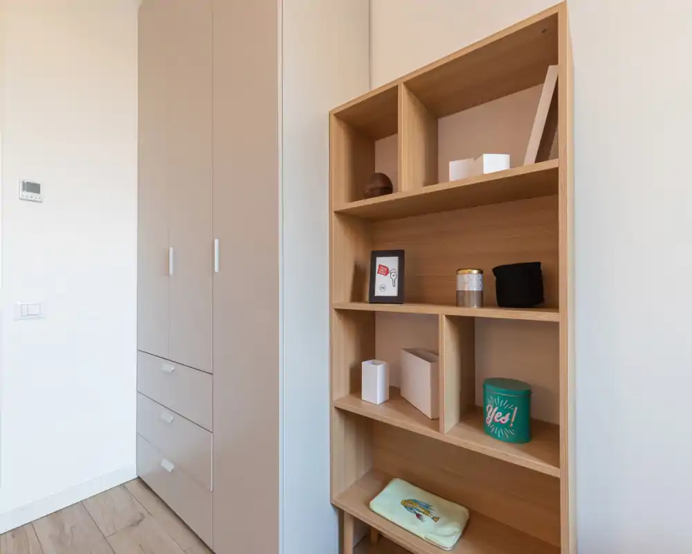 Bright, minimal bedroom corner with built-in wardrobe and wooden shelving holding decorative boxes and small accents.