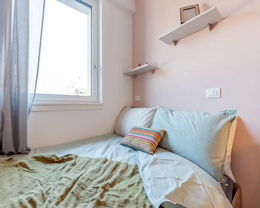 Sunny small bedroom with a neatly made bed, pastel walls and natural light from a large window — cozy, airy and well staged for short-term rentals.