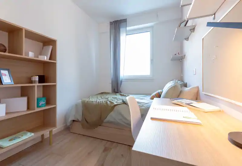 Bright, compact bedroom with a single bed, study desk and shelving — ideal for a student or solo occupant. Natural light from the window and warm desk lighting create a pleasant, functional space.