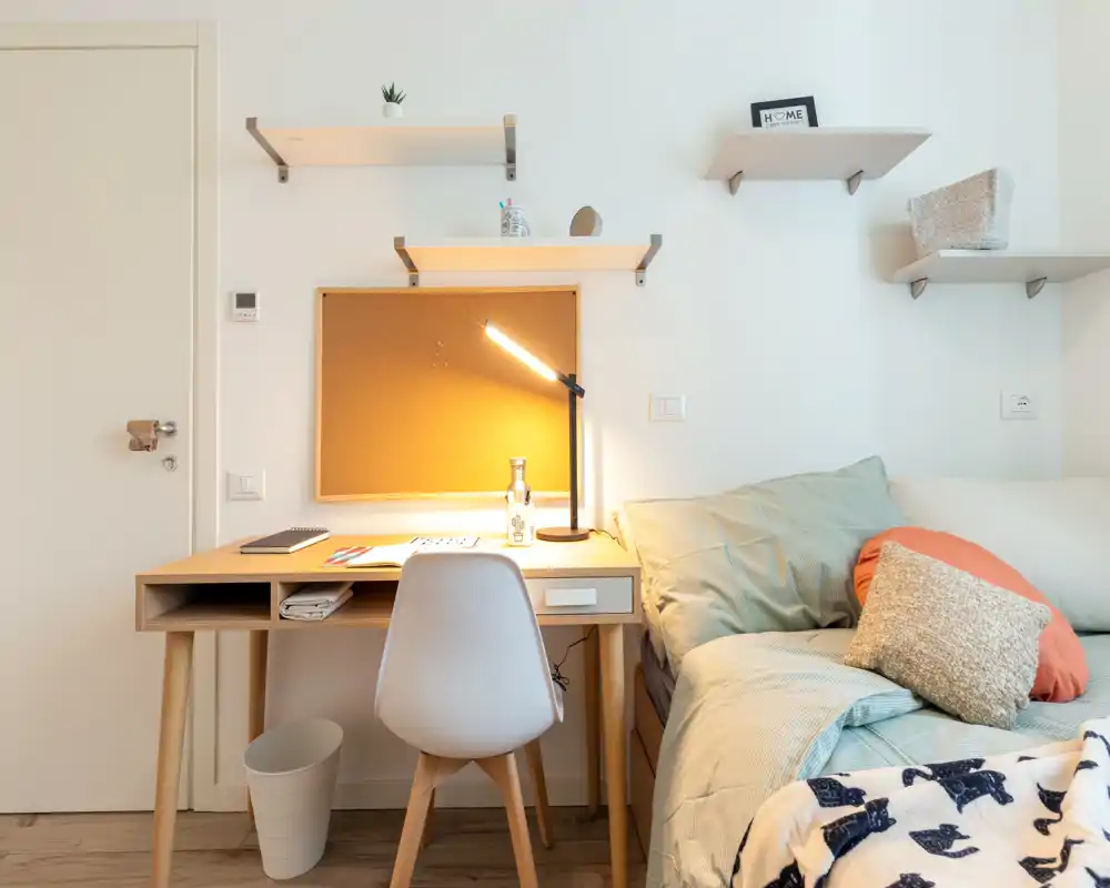 Bright, cozy bedroom corner with a made bed, bedside pillows and a wooden study desk lit by a warm desk lamp — ideal for a student or home office setup.
