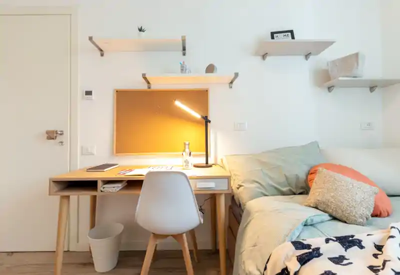 Bright, cozy bedroom corner with a made bed, bedside pillows and a wooden study desk lit by a warm desk lamp — ideal for a student or home office setup.