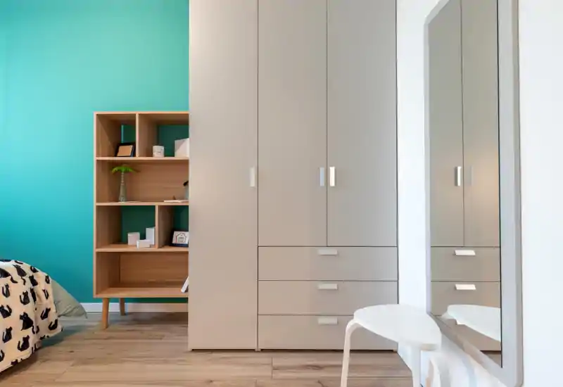 Bright, modern bedroom corner with a large wardrobe, shelving unit and a partial view of the bed with patterned bedding. Clean lines, wood flooring and a teal accent wall give a fresh, contemporary look.
