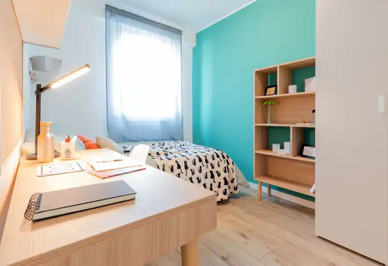 Bright, compact bedroom with a single bed, study desk in the foreground and a turquoise accent wall. The space is clean, softly lit by a window and furnished for a student or single occupant.