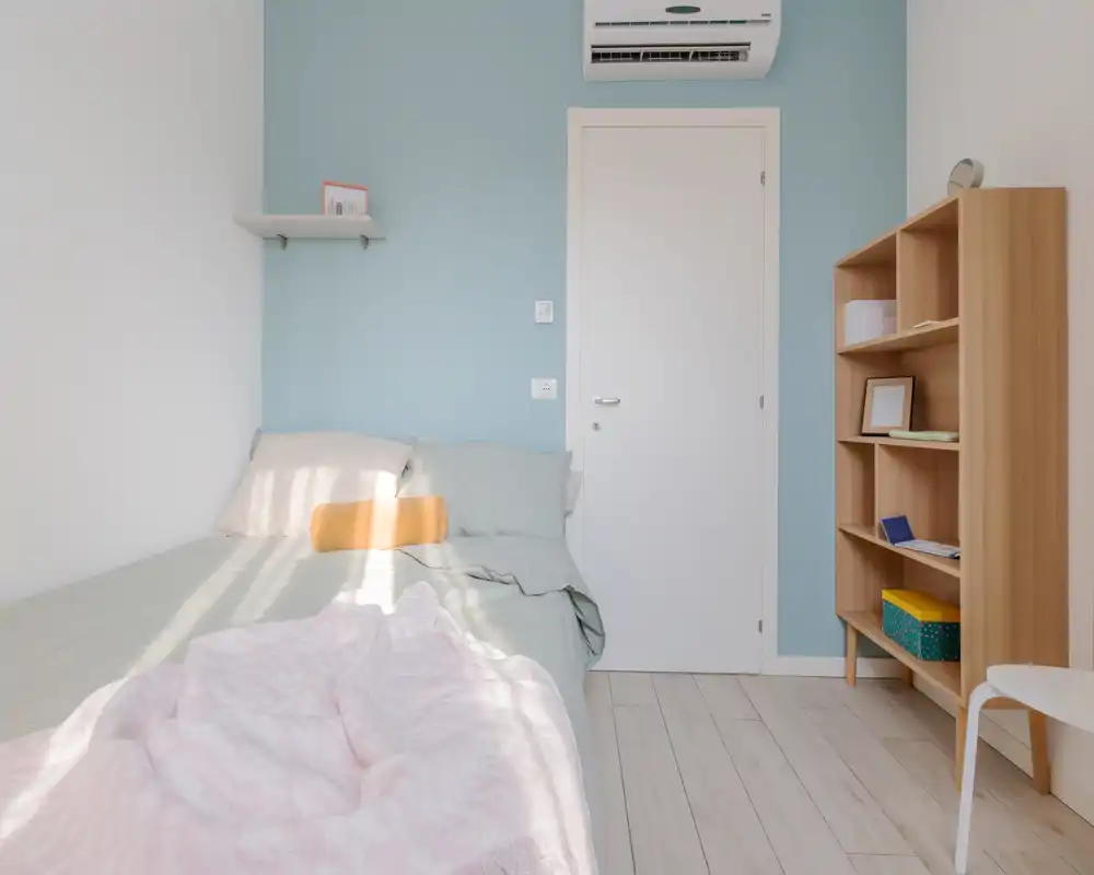 Bright, minimalist bedroom with a comfortable bed, soft pastel linens, a wooden shelving unit and natural light creating a calm atmosphere.