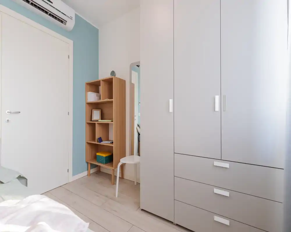 Small, bright bedroom corner with a wardrobe, wooden shelving and a glimpse of the bed. Neutral palette and natural light create a calm, tidy atmosphere suitable for rental listings.
