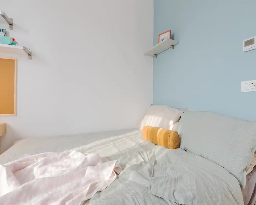 Bright, softly styled bedroom with a large bed dressed in pale linens, decorative bolster pillow and a light throw. Natural light highlights the calming pastel wall and tidy shelving — ideal for rental photos showcasing comfort.
