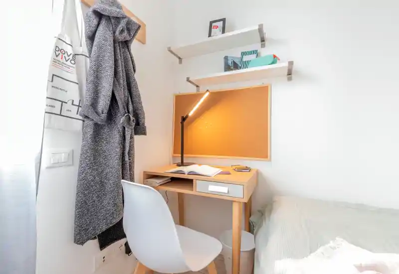 Compact bedroom corner with a small wooden desk, chair and bedside area; bright natural light, neat bedding and a reading lamp create a cozy, functional space ideal for studying or relaxing.