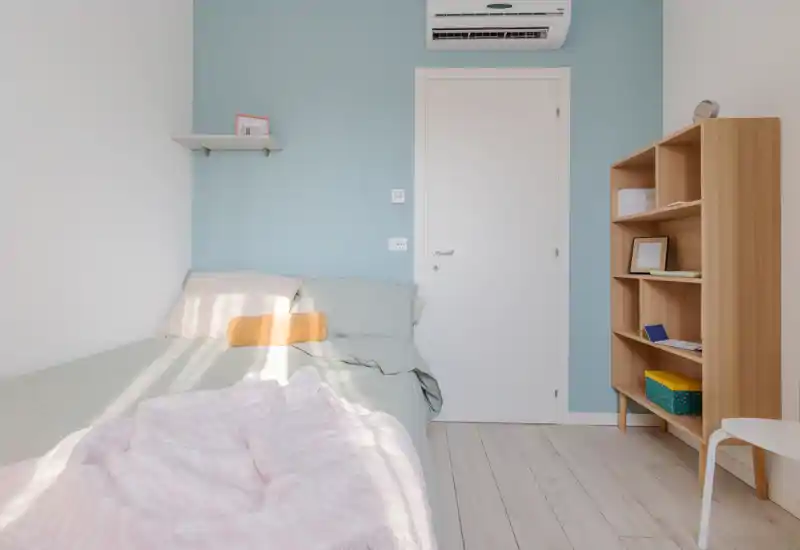 Bright, minimalist bedroom with a comfortable bed, soft pastel linens, a wooden shelving unit and natural light creating a calm atmosphere.