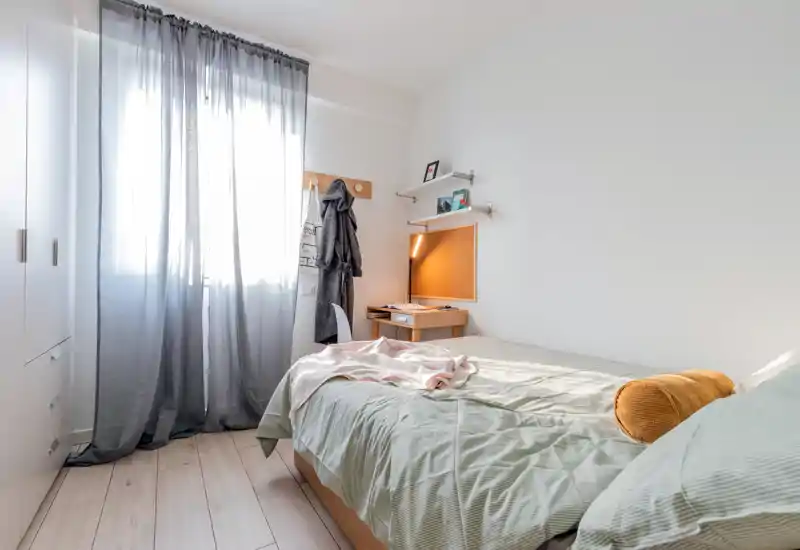 Bright, compact bedroom with a neatly made single bed, soft green bedding, warm accent pillow and a small desk with a reading lamp by the wall.