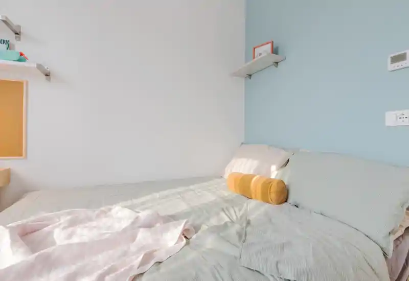 Bright, softly styled bedroom with a large bed dressed in pale linens, decorative bolster pillow and a light throw. Natural light highlights the calming pastel wall and tidy shelving — ideal for rental photos showcasing comfort.