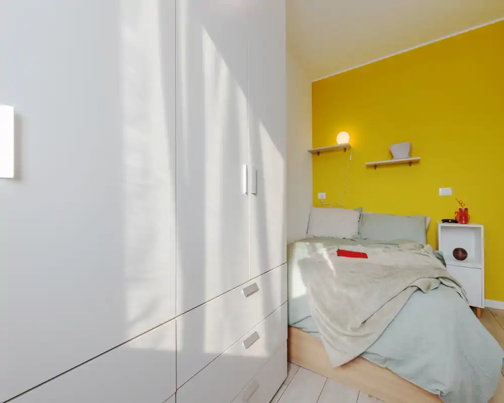 Camera da letto moderna e luminosa con letto matrimoniale davanti a una parete gialla, biancheria morbida, una lampada su mensola e un armadio bianco con spazio contenitivo.
