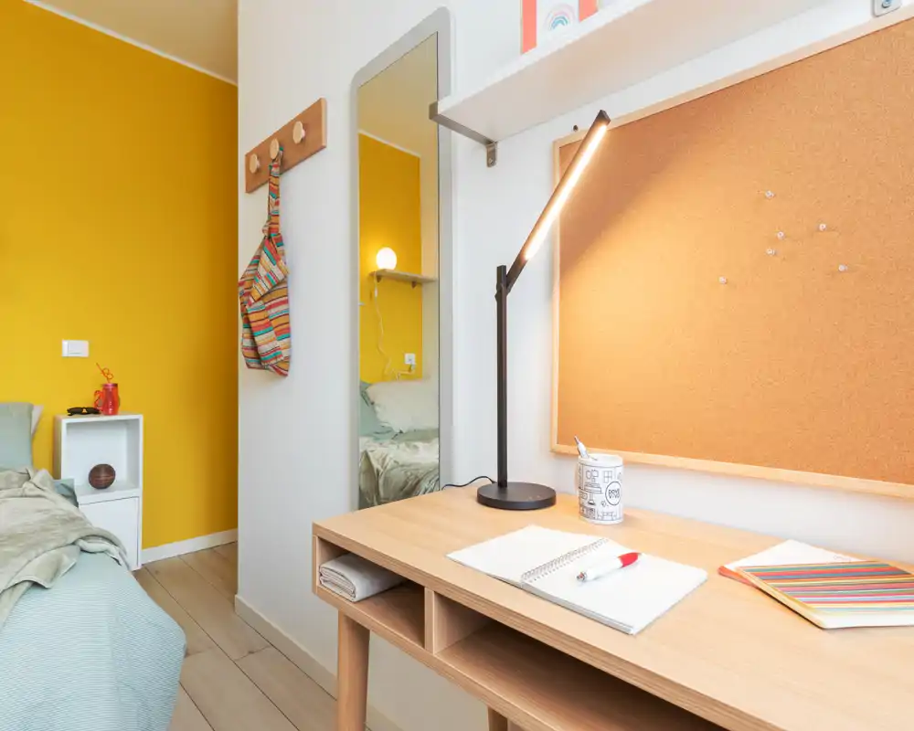 Angolo camera luminoso e moderno con scrivania in legno ordinata, lampada da tavolo e bacheca in sughero; il letto con biancheria morbida e la parete gialla creano uno spazio accogliente per dormire e studiare.