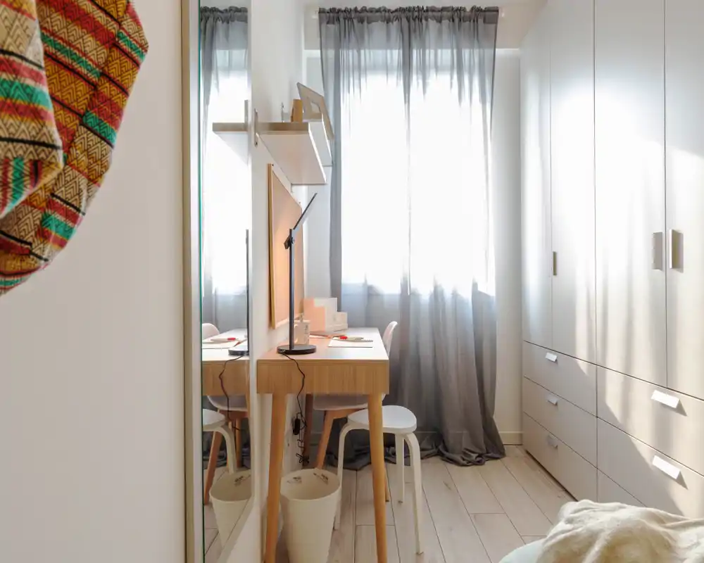 Angolo camera luminoso e compatto con scrivania in legno, armadio e una piccola porzione del letto visibile; tende morbide diffondono la luce naturale creando un accogliente angolo per lavoro e riposo.