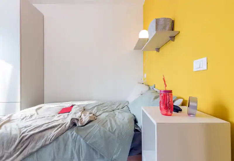 Camera da letto luminosa e moderna con letto comodo, comodino e una parete d'accento gialla; biancheria morbida e una mensola a muro creano un ambiente accogliente e ben illuminato.