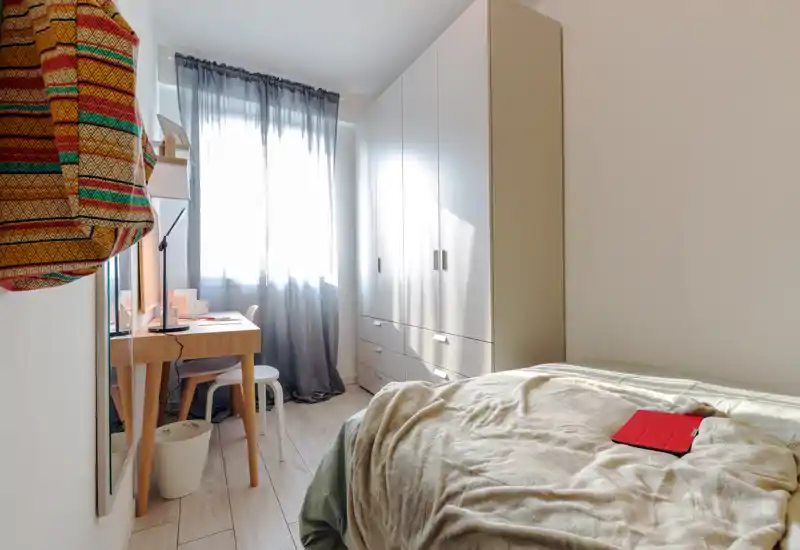 Camera da letto luminosa e ben allestita con letto avvolto in coperte morbide, un armadio e una piccola scrivania vicino alla finestra che lascia entrare molta luce naturale.