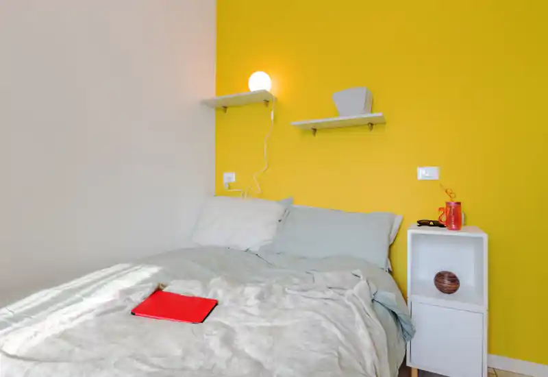 Angolo notte luminoso e accogliente con letto matrimoniale contro una parete gialla, mensole e un piccolo comodino bianco — perfetto per annunci che evidenziano calore e luce naturale.