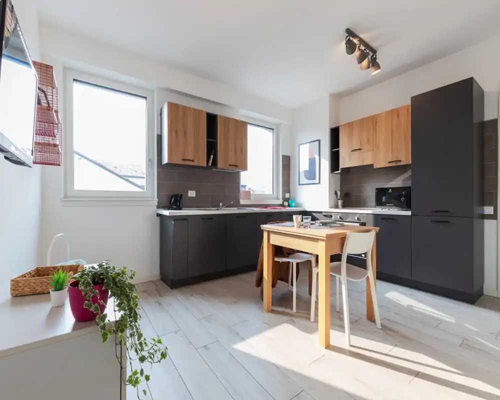 Bright modern kitchen with dark lower cabinets and warm wood upper units, a small dining table in the center, and large windows providing abundant natural light — a practical, well-styled cooking and dining space.
