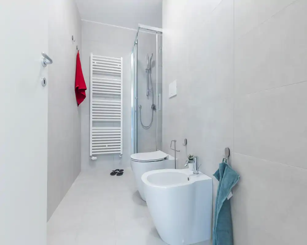 Contemporary, bright bathroom with a wall-mounted toilet, bidet, glass-enclosed shower and heated towel rack. Clean minimalist tiles and small decorative towel details create a fresh, modern look.