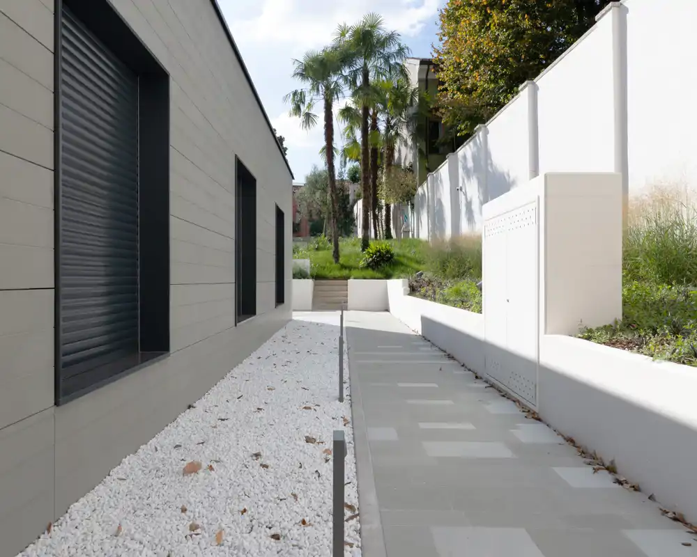 Modern exterior walkway with paved path, white gravel bed and landscaped greenery, framed by a building façade and retaining wall.