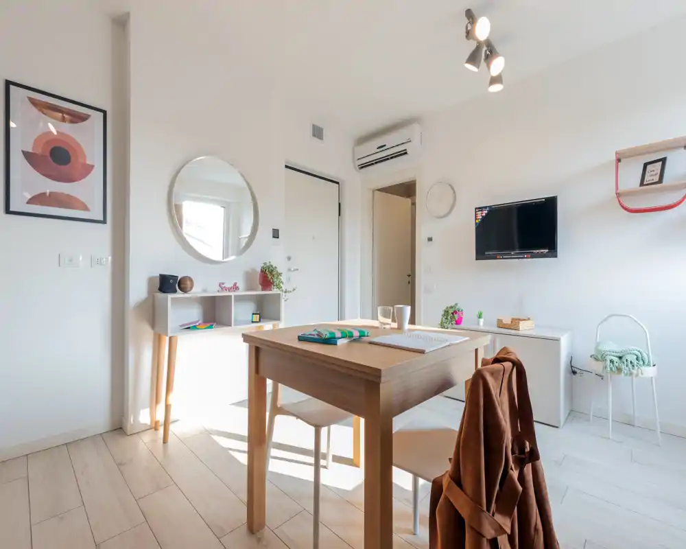 Bright, modern dining area with a wooden table set for two, minimalist shelving and soft decorative touches — ideal for a compact apartment.