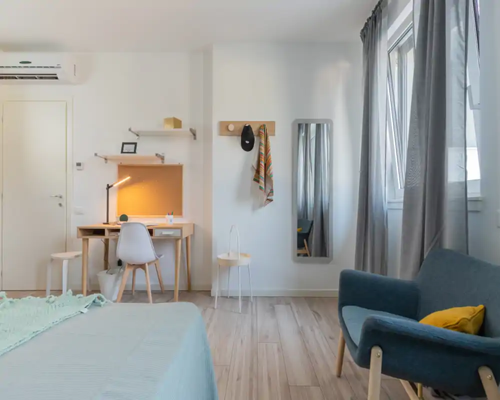 Bright, modern bedroom with a comfortable bed in the foreground, a desk with warm task lighting, a full-length mirror and a cozy armchair by the window.