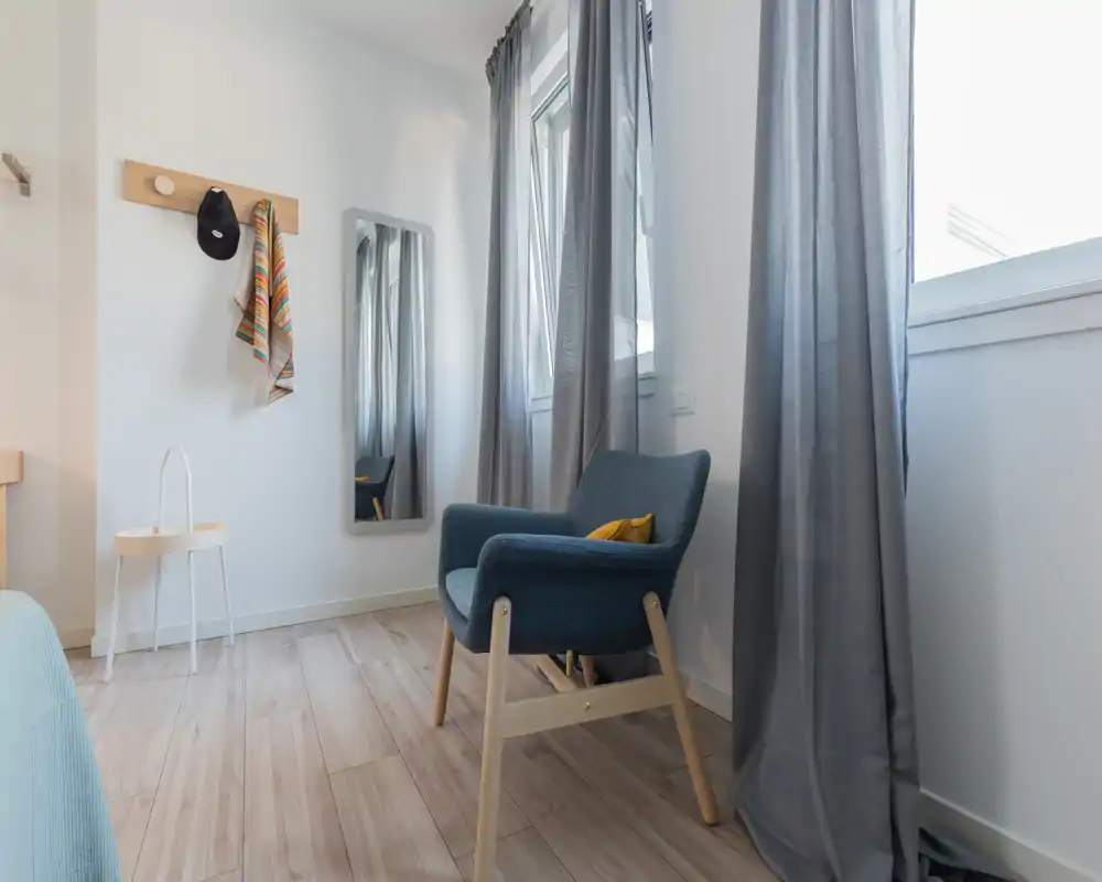Bright, minimalist bedroom corner with a cushioned armchair, full-length mirror, and soft grey curtains; natural light fills the space for a calm, modern feel.
