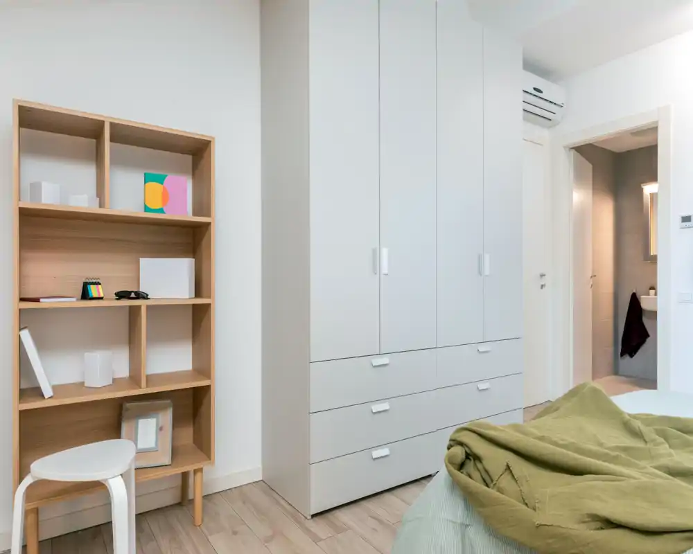Bright, modern bedroom corner with a visible bed, large white wardrobe and wooden shelving; calm neutral tones and soft textiles create a welcoming atmosphere ideal for listings.