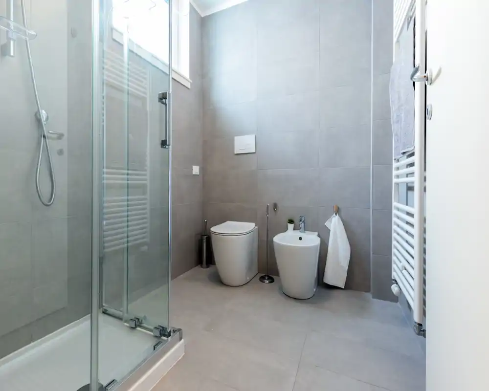 Modern, light bathroom with a walk-in shower, wall-mounted toilet, bidet and towel radiator; neutral grey tiles and bright natural light create a clean, minimalist look.
