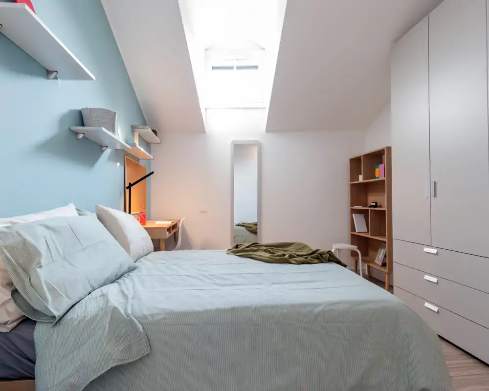 Bright, modern bedroom with a neatly made double bed under a skylight, soft blue accent wall, built-in wardrobe and a small desk — ideal for a cozy, well-lit rental.